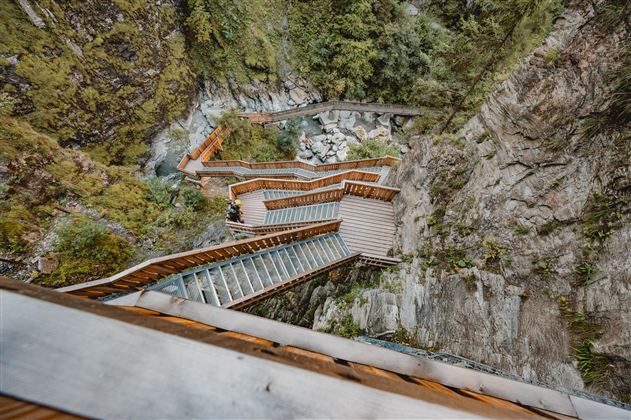 A wooden staircase winds through an impressive rock landscape. Below, a small stream flows between the stones.
