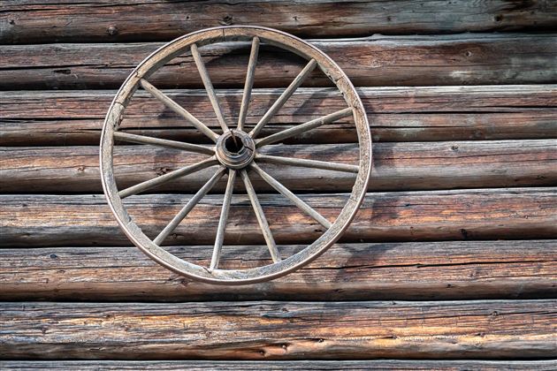 An old wooden wheel hangs on a rustic wooden wall. The structure of the wall emphasizes the vintage aesthetic.