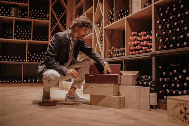 Ein Mann sitzt in einem Weinkeller und schaut in eine Kiste. Um ihn herum sind zahlreiche Weinflaschen in Regalen aufgestellt.