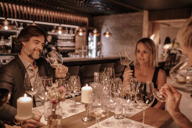 Eine elegante Dinner-Atmosphäre mit einem romantischen Ambiente. Zwei Personen genießen Wein und interagieren am Tisch mit Kerzenlicht und vielen Weingläsern.