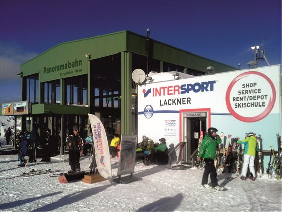 A ski resort with the panoramic lift and an Intersport shop. Many skiers and snowboarders are nearby on the snow.