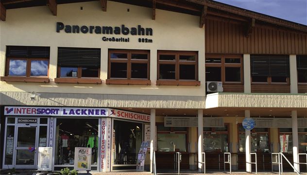 A building with a sign "Panoramabahn" and shops underneath. There are large windows and an inviting entrance area.