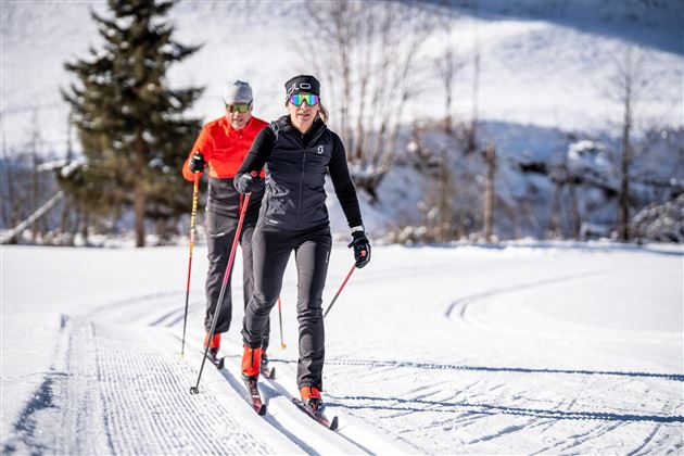 Zwei Frauen machen Langlauf in einer verschneiten Landschaft. Im Hintergrund sind Bäume und schneebedeckte Hügel zu sehen.