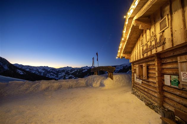 Eine rustikale Berghütte in der verschneiten Landschaft bei Dämmerungslicht. Die Umgebung ist von den schneebedeckten Bergen umgeben, und der Himmel hat einen schönen Farbverlauf.