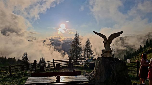 A picturesque mountain landscape with mist and sun rays breaking through the clouds. In the foreground stands a statue of an eagle and there are trees as well as a bench.