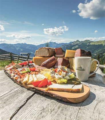 A wooden board with a selection of cheese varieties and sausage, surrounded by mountains and a blue sky. In the foreground, there is a cup on a rustic table.