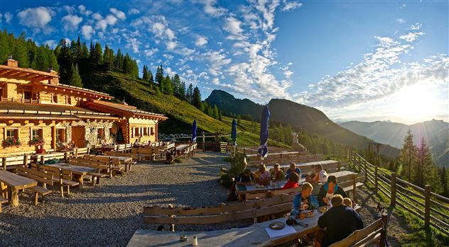 A cozy mountain cabin with a wooden terrace and outdoor seating. In the background, green mountains and a clear sky can be seen.