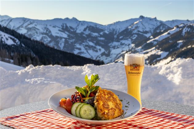 Ein Teller mit einer köstlichen Speise und frischem Gemüse steht auf einem Tisch. Im Hintergrund sind schneebedeckte Berge und ein Bierglas zu sehen.