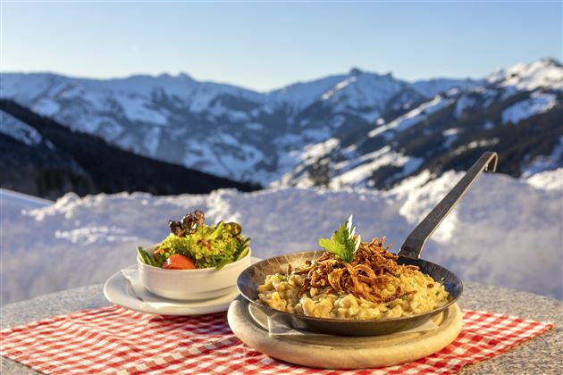 Ein herzhaftes Gericht in einer Pfanne, garniert mit Zwiebeln, neben einem frischen Salat. Im Hintergrund sind schneebedeckte Berge und ein klarer Himmel zu sehen.