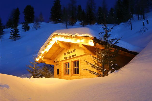 Eine gemütliche Almhütte im Schnee, beleuchtet von warmem Licht. Umgeben von schneebedeckten Bäumen und einer nächtlichen Winterlandschaft.