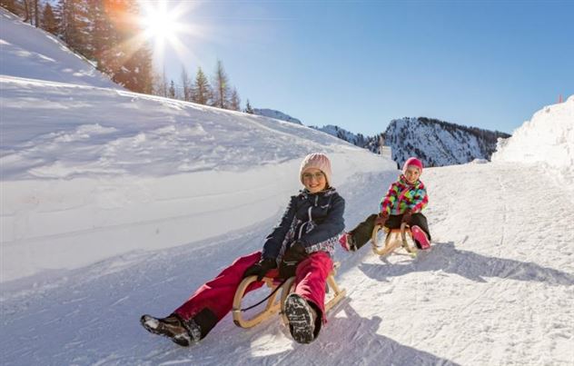 Zwei Kinder fahren auf Schlitten in einer winterlichen Landschaft. Der Himmel ist blau und die Sonne scheint hell.