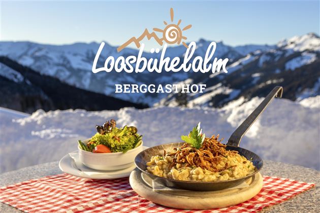 Ein leckerer Teller mit traditionellem Essen und frischem Salat vor einer beeindruckenden Bergkulisse. Im Hintergrund Schnee und Berge, die die alpine Atmosphäre unterstreichen.