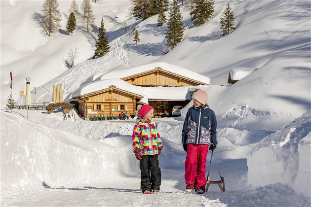 Zwei Kinder stehen im Schnee vor einer Holzschutzhütte. Die Umgebung ist verschneit, umgeben von Bergen und Bäumen.