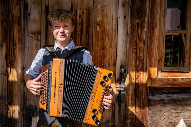 Ein junger Mann steht vor einer Holzwand und spielt ein Akkordeon. Er trägt traditionelle Kleidung und lächelt freundlich.