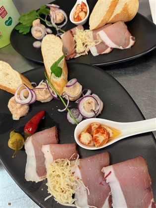 Ein Teller mit verschiedenen Wurstsorten, frischem Brot und dekorativen Zutaten wie Zwiebeln und Kräutern. Dazu kleine Schalen mit schmackhaften Beilagen.