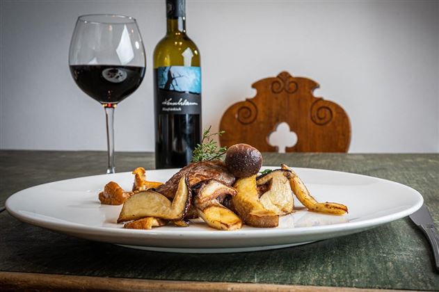Ein köstliches Gericht mit Pilzen und Bratkartoffeln auf einem großen weißen Teller. Im Hintergrund steht ein Glas Rotwein und eine Flasche Wein.