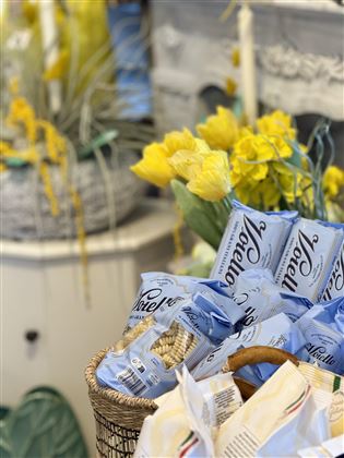 Ein Korb mit blauen Verpackungen von Snacks steht vor gelben Blumen. Die Komposition wirkt einladend und farbenfroh.