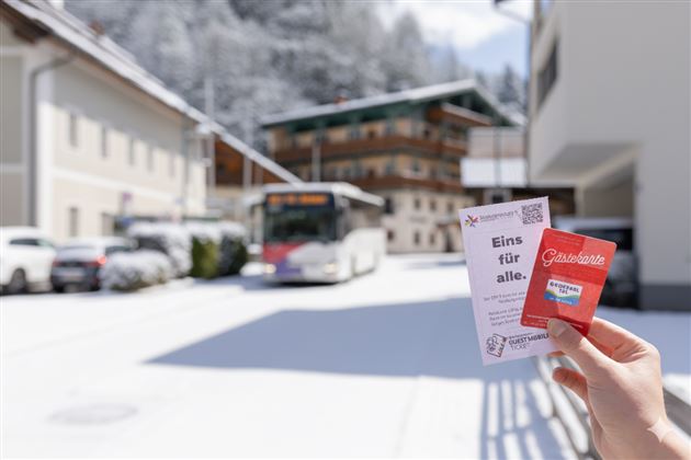 A view of a snow-covered street with a bus and several buildings in the background. Someone is holding two information cards in their hand.