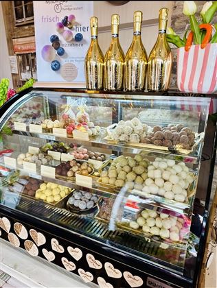 Ein Kühlschrank voller leckerer Pralinen in verschiedenen Geschmacksrichtungen. Oben stehen Flaschen mit Sekt und frische Blumen in einem Topf.