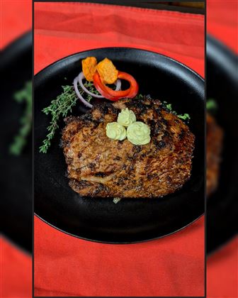 Ein saftiges Stück Fleisch auf einem schwarzen Teller, garniert mit frischen Kräutern und Gemüse. Im Hintergrund liegt ein rotes Tuch.