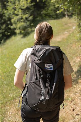 A person with a backpack is standing on a hiking trail. In the background, trees and green grass are visible.