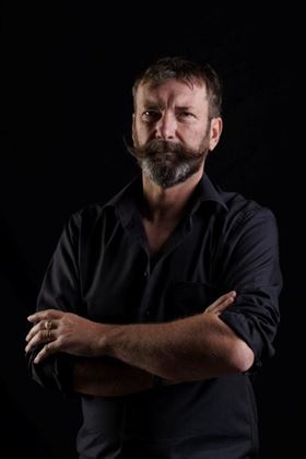 A man with a distinctive beard stands in a dark environment. He is wearing a black shirt and has a serious expression on his face.