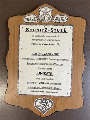 Ein Holzschild mit dem Titel "Schnitz-Stube". Es enthält Informationen über eine Werkstatt für handgefertigte Holzschnitzarbeiten.
