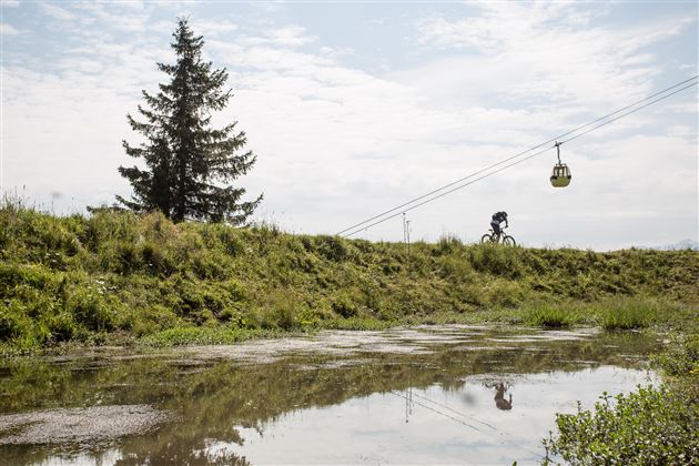 Eine grüne Wiese mit einem Fahrradfahrer und einer Seilbahn im Hintergrund. Im Vordergrund spiegelt sich der Himmel im flachen Wasser.