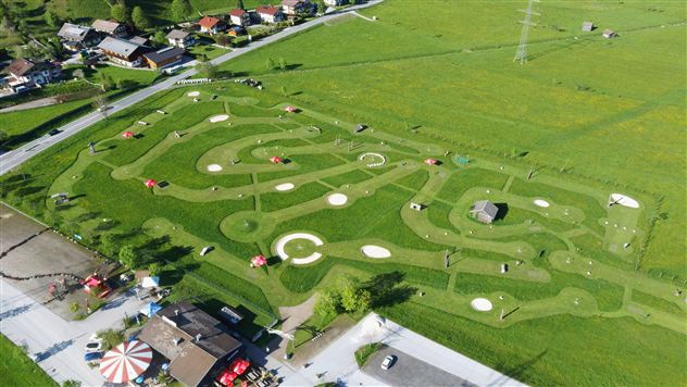 A large, green meadow with an exciting go-kart track designed in curving lines. In the background, houses and a small amusement area can be seen.