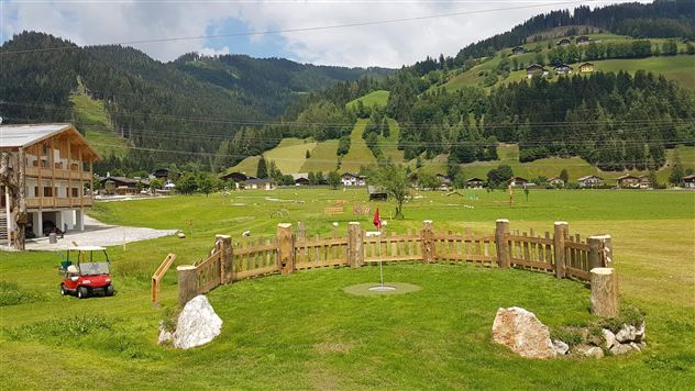 A picturesque landscape with green meadows and gentle hills. In the foreground, there is a wooden fence with some stones as well as a golf cart.