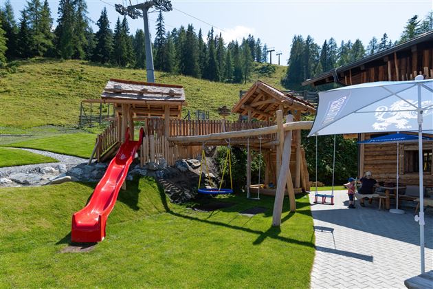 A playground with a red slide, swings, and wooden structures. In the background, green meadows and trees can be seen.