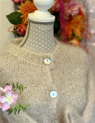 Ein weicher, beiger Cardigan mit Knöpfen auf einem Kleiderständer. Im Hintergrund sind bunte Blumen sichtbar.