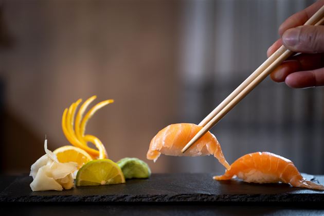 Ein kunstvoll angerichtetes Sushi-Plateau mit Lachs, Ingwer und Limette. Ein Hand mit Essstäbchen hebt ein Stück Sushi.