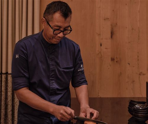 Ein Koch schneidet sorgfältig frische Zutaten in einer modernen Küche. Der Raum hat eine warme Holzverkleidung und eine elegante Atmosphäre.