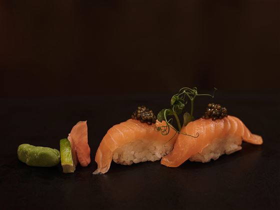 Zwei Stücke Sushi mit Lachs, garniert mit Kaviar und frischem Gemüse. Daneben liegt Wasabi und etwas eingelegter Ingwer.