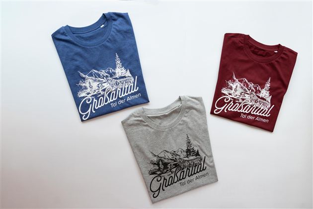 Three T-shirts in different colors are lying on a white background. Each shirt features a colorful design with a mountain motif and the inscription “Grasaital – Valley of the Pastures.”