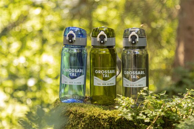 Three water bottles in different colors are standing on a moss-covered stone in the forest. The bottles are printed with the inscription "GROSSARLTAL".