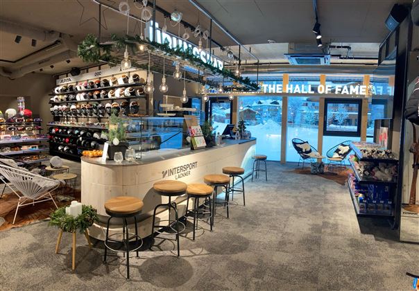 Ein moderner Innenraum mit einem Verkaufsbereich und einer gemütlichen Theke. Regale mit verschiedenen Artikeln sind sichtbar, und große Fenster bieten einen Blick nach draußen.