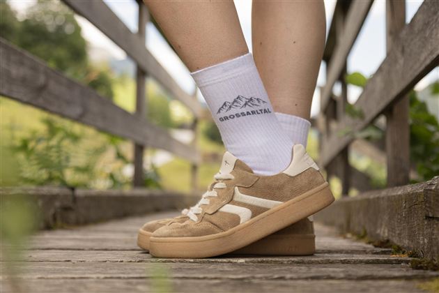 Comfortable sneakers are on a wooden walkway. In the background, green meadows and trees are visible.