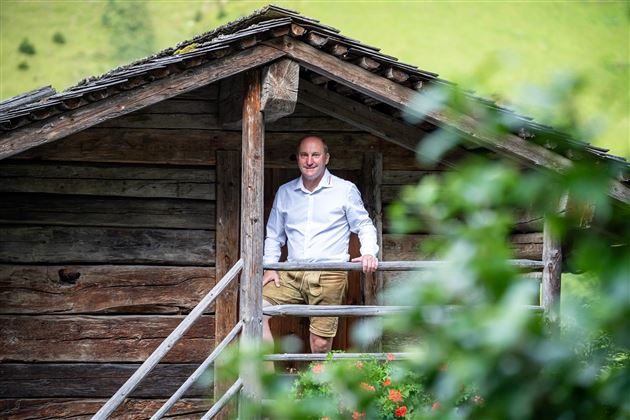 Ein Mann steht auf dem Balkon einer rustikalen Hütte. Im Hintergrund sind grüne Wiesen und Bäume zu sehen.