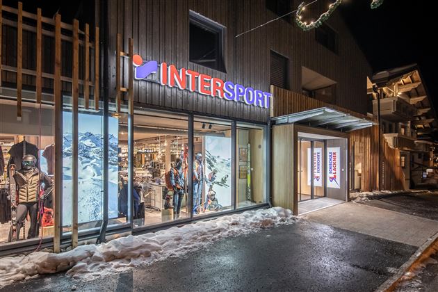 Ein Intersport-Geschäft bei Nacht, beleuchtet und einladend. Der Weg ist schneebedeckt und das Gebäude hat eine moderne Architektur.