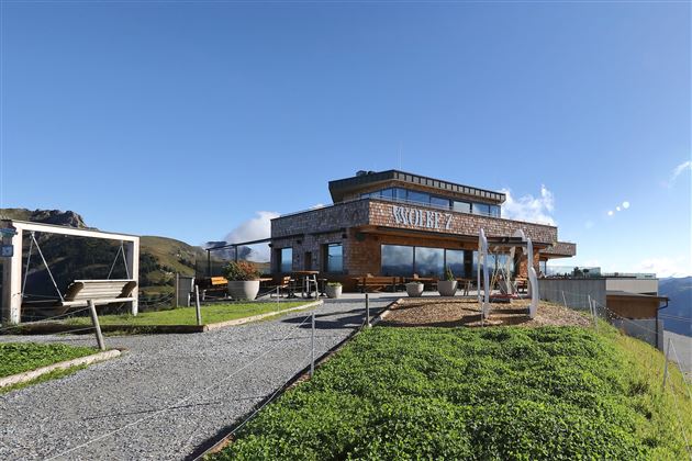 A modern building on a mountain with a clear blue sky and lots of greenery in the background. The area around the building is inviting with outdoor seating options.
