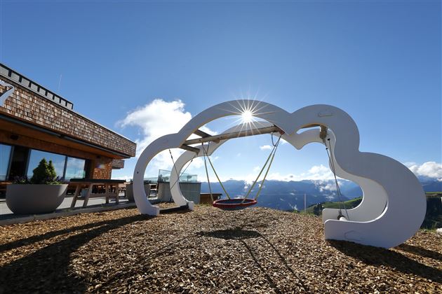 A modern swing set in cloud shape with a view of the mountains. The sky is clear and the sun is shining.