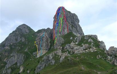 Eine große Felswand mit mehreren Kletterrouten in verschiedenen Farben. Die Umgebung ist grün und bergig mit bewölktem Himmel.