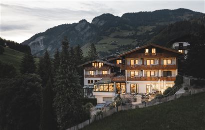 Ein charmantes Berghotel in der Dämmerung, umgeben von hohen Bäumen. Die Lichter im Hotel sind warm und einladend.