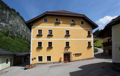 Ein gelbes Gebäude mit mehreren Fenstern und einem einladenden Eingang. Im Hintergrund sind grüne Berge und eine sonnige Atmosphäre zu sehen.