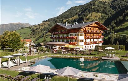 A beautifully designed hotel with wood paneling is located in a picturesque mountain landscape. In the foreground, there is an inviting pool with loungers and sun umbrellas.