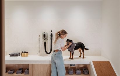Eine Frau trocknet einen Hund mit einem Handtuch in einem modernen Raum. Im Hintergrund sind Regale mit Handtüchern sichtbar.
