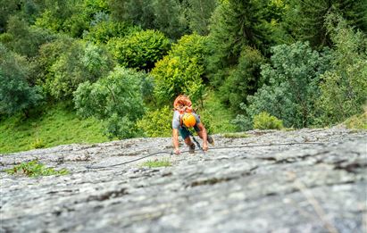Eine Person klettert an einer steilen Felswand. Im Hintergrund sind grüne Bäume und eine üppige Landschaft zu sehen.