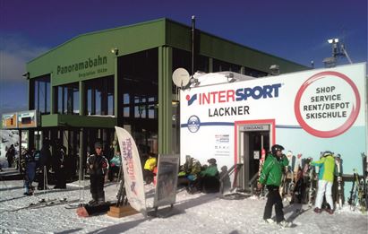 Ein Skigebiet mit der Panoramabahn und einem Intersport-Shop. Viele Skifahrer und Snowboarder sind in der Nähe auf dem Schnee.